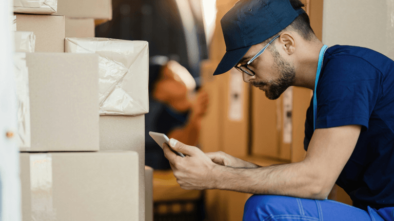This image shows a delivery or warehouse worker sitting on the ground next to stacked cardboard boxes, looking at their phone. The worker is wearing a navy blue uniform, a cap, and glasses. It seems like they may be checking details or coordinating tasks while surrounded by packages in a logistics environment. In the background, another individual is partially visible, suggesting a busy and active workplace