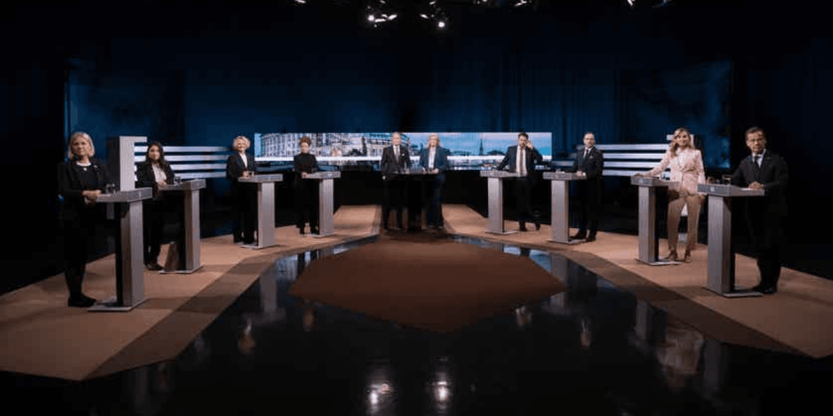 Eight individuals standing at podiums in a debate setting with a dark background