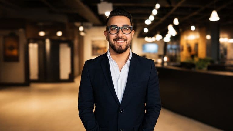Amirali Adib standing in a modern office wearing a navy suit and glasses.