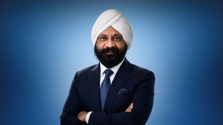A man wearing a white turban and dark suit with a blue tie stands confidently against a gradient blue background, arms crossed and smiling slightly.
