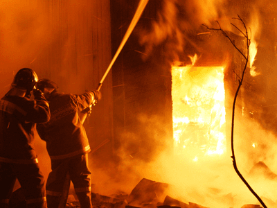 Two firefighters direct a hose at a structure engulfed in flames during an intense nighttime fire.