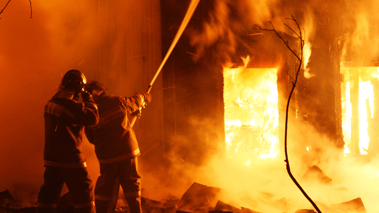 Two firefighters direct a hose at a structure engulfed in flames during an intense nighttime fire.