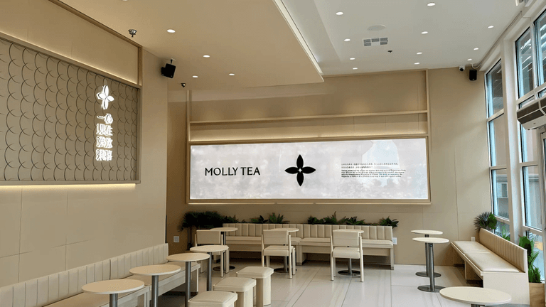 Interior of a modern Molly Tea location, showcasing minimalist decor with light beige walls, sleek furniture, and ambient lighting. The store features a large wall with the brand’s logo, 'MOLLY TEA,' alongside a digital display with messaging in both English and Chinese, reflecting the cultural blend of the business. The clean and contemporary design reflects the modern yet culturally rooted identity of the brand, which has gained traction with the help of Chowbus technology solutions.