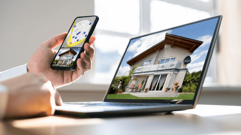 Person holding smartphone displaying a house image while using a laptop with the same view
