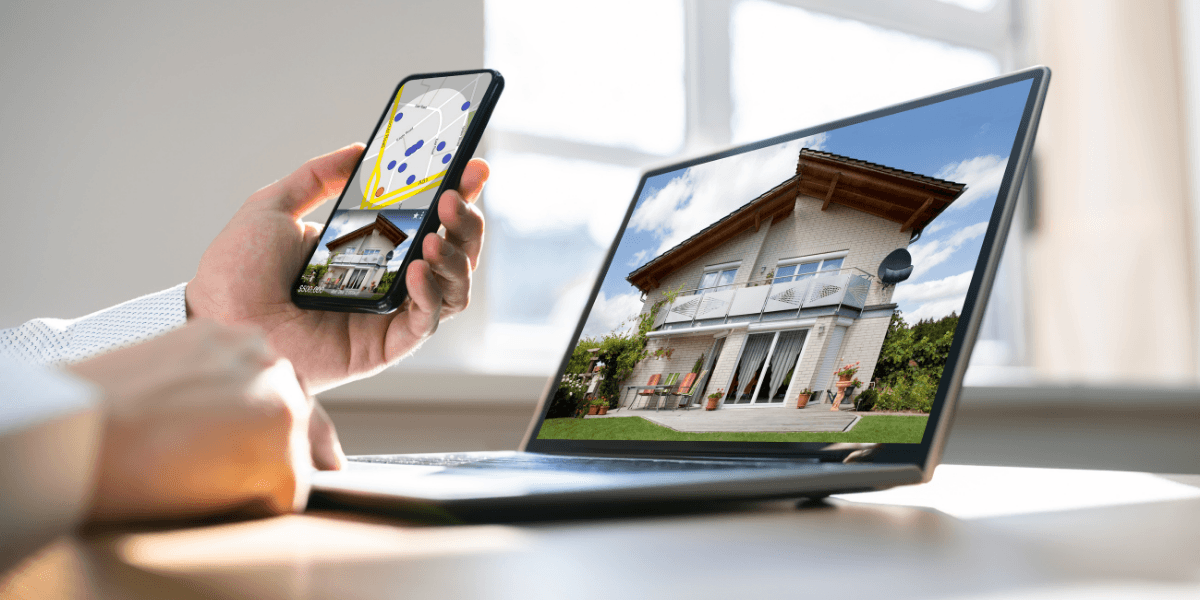 Person holding smartphone displaying a house image while using a laptop with the same view