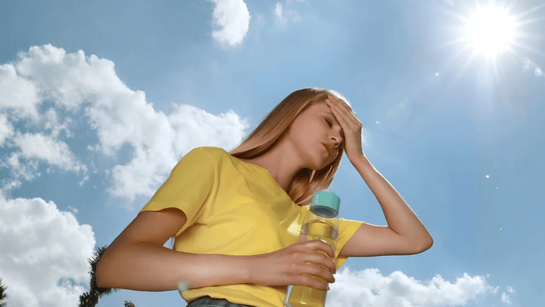 A woman in a yellow shirt holds a water bottle, shielding her eyes from the sun