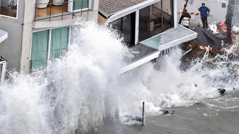 Waves crash against a building, causing damage and flooding near the water's edge
