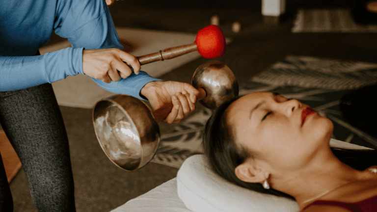 Sound healing practitioner playing singing bowls beside a relaxed participant during a wellness session.