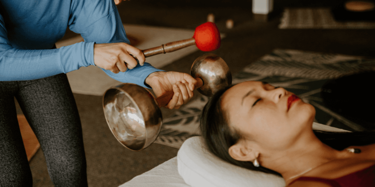 Sound healing practitioner playing singing bowls beside a relaxed participant during a wellness session.