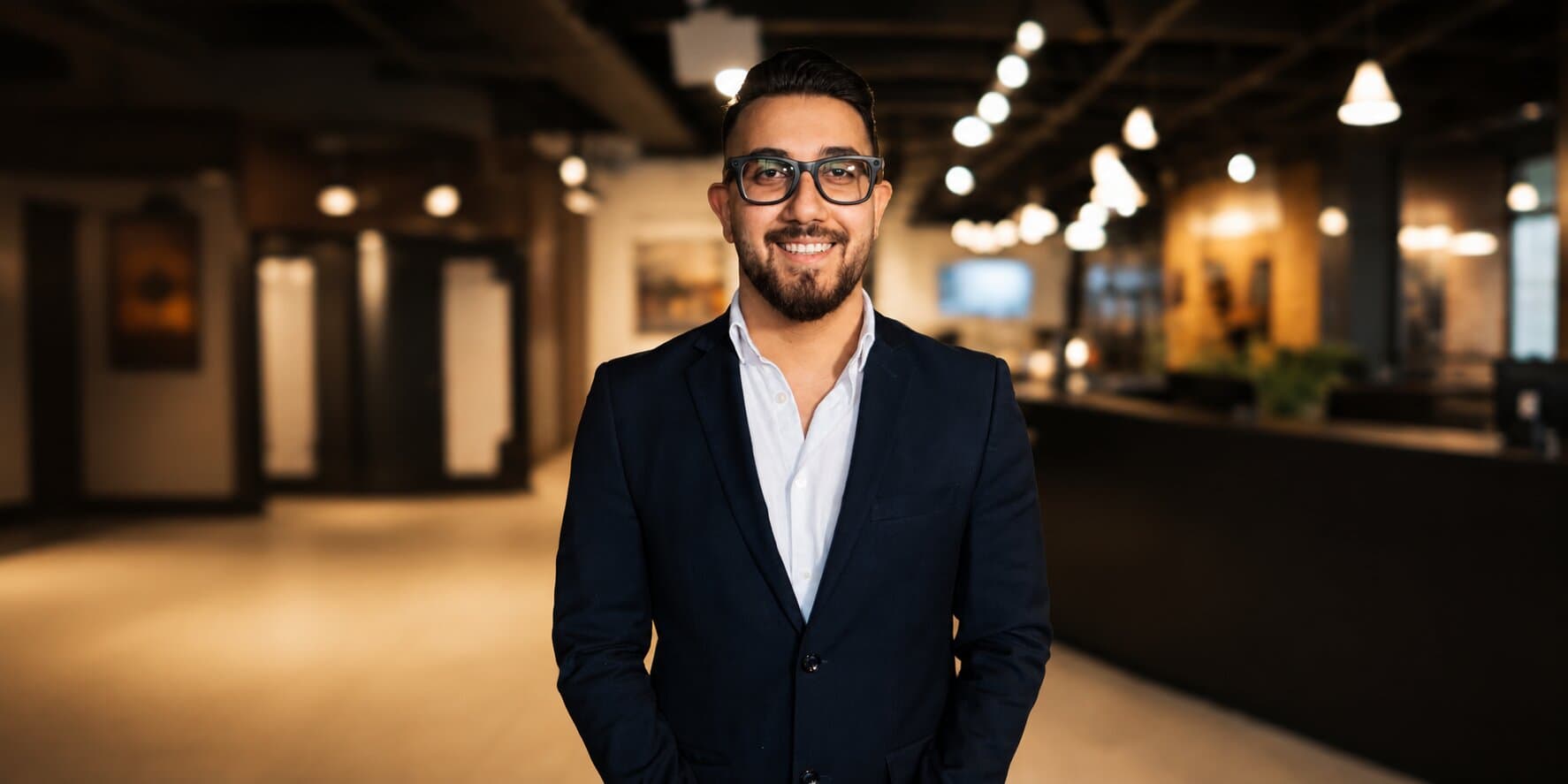 Amirali Adib standing in a modern office wearing a navy suit and glasses.