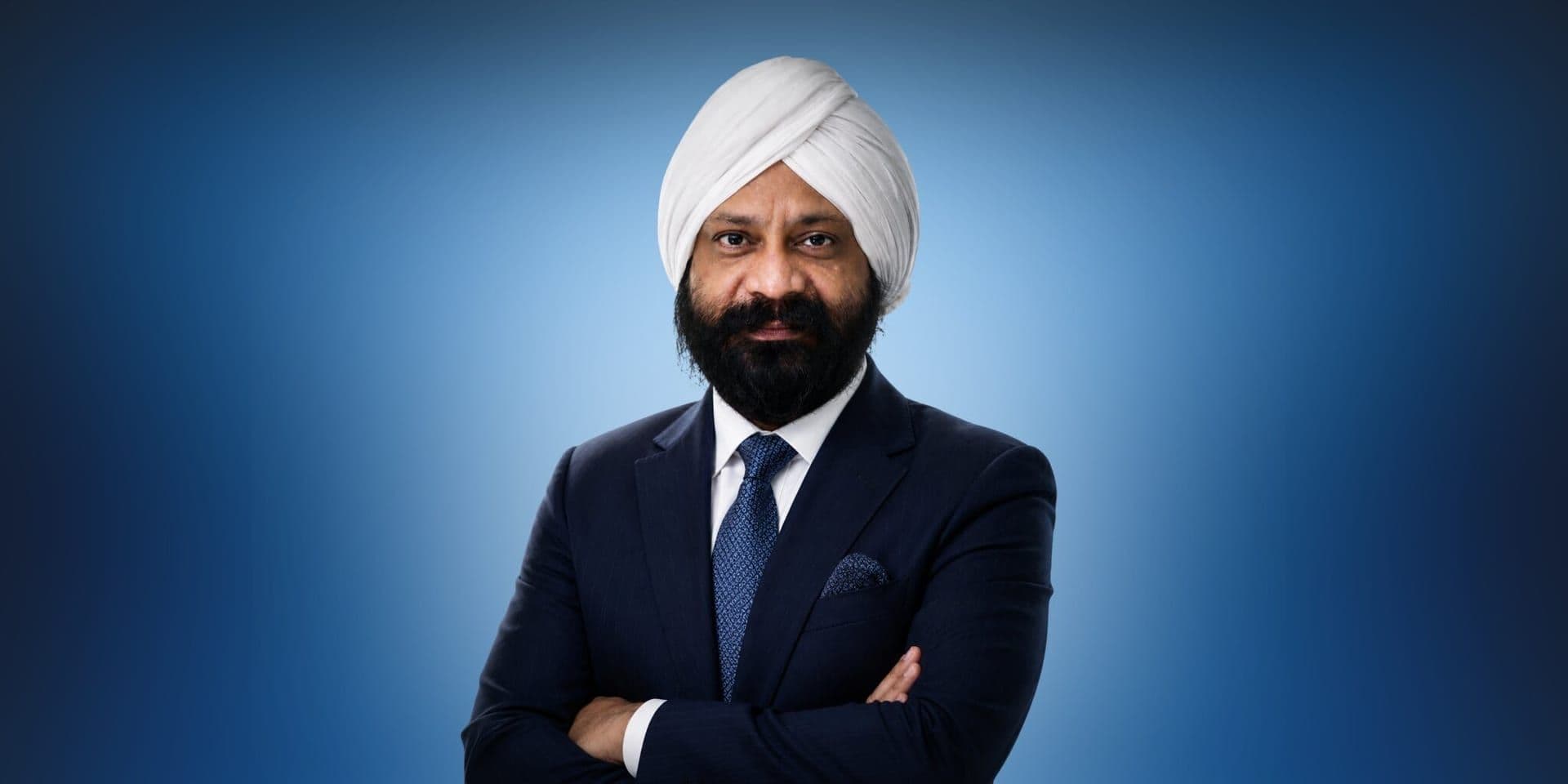 A man wearing a white turban and dark suit with a blue tie stands confidently against a gradient blue background, arms crossed and smiling slightly.