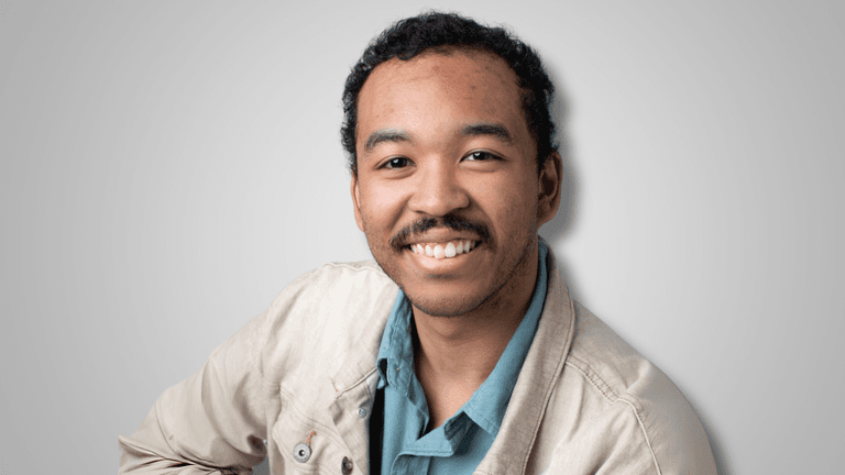 Professional headshot of a young man smiling at the camera, wearing a light beige jacket over a teal shirt, with a neutral gray background.
