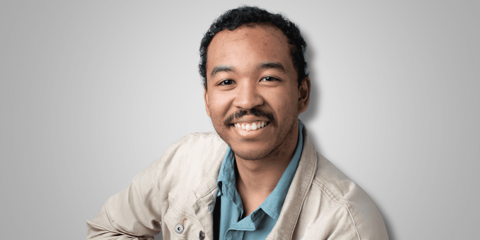 Professional headshot of a young man smiling at the camera, wearing a light beige jacket over a teal shirt, with a neutral gray background.