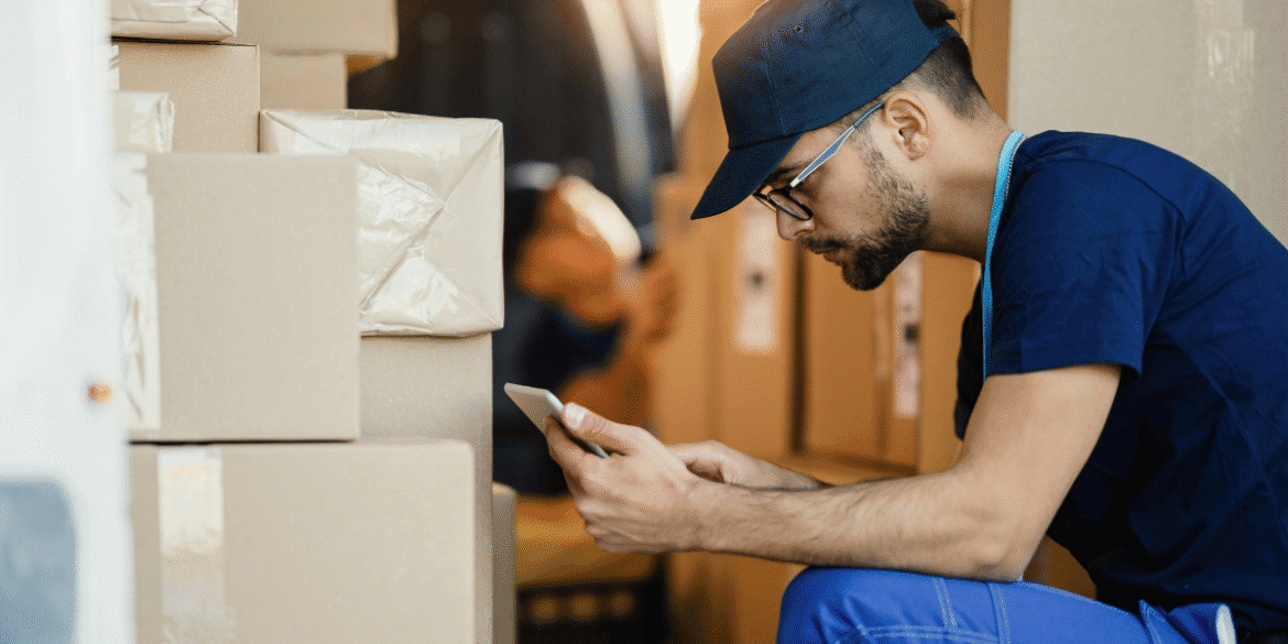 This image shows a delivery or warehouse worker sitting on the ground next to stacked cardboard boxes, looking at their phone. The worker is wearing a navy blue uniform, a cap, and glasses. It seems like they may be checking details or coordinating tasks while surrounded by packages in a logistics environment. In the background, another individual is partially visible, suggesting a busy and active workplace