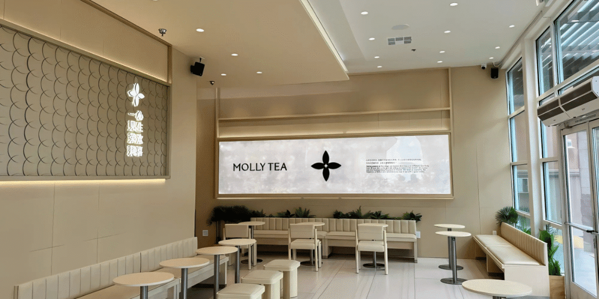 Interior of a modern Molly Tea location, showcasing minimalist decor with light beige walls, sleek furniture, and ambient lighting. The store features a large wall with the brand’s logo, 'MOLLY TEA,' alongside a digital display with messaging in both English and Chinese, reflecting the cultural blend of the business. The clean and contemporary design reflects the modern yet culturally rooted identity of the brand, which has gained traction with the help of Chowbus technology solutions.