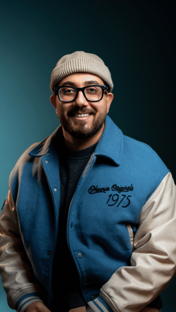 Amirali Adib smiling in a blue varsity jacket, glasses, and beige beanie against a dark studio background.