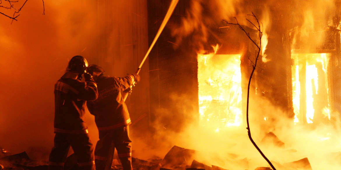 Two firefighters direct a hose at a structure engulfed in flames during an intense nighttime fire.